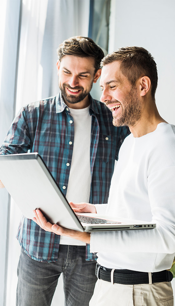 Two smiling young businessmen using laptop, standing close to a large window at an office. Light shines through.