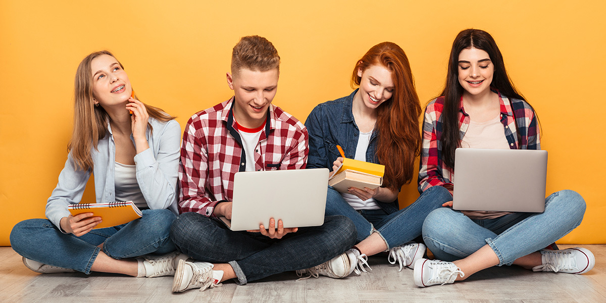 Four young students sitting on the floor with laptops and notepads